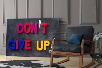 Black glass wall art with colorful 3D letters spelling "DON'T GIVE UP" displayed in modern interior beside wooden chair.