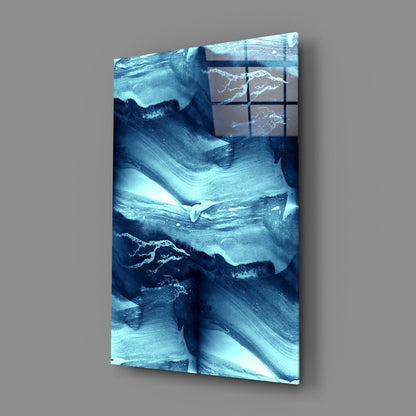 Angled view of Blue Serenity glass wall art displaying layered blue and white abstract patterns with a 3x3 grid overlay on gray background.