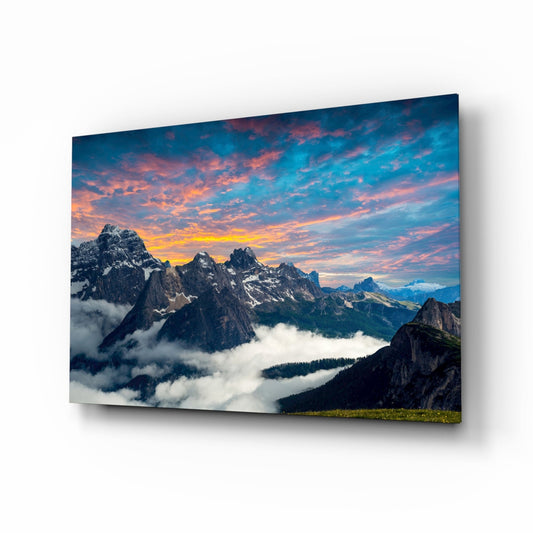 Glass wall art displaying dramatic mountain peaks with clouds below, fiery orange and pink sunset sky above, photographed at an angle showing the panel's depth and shadow.