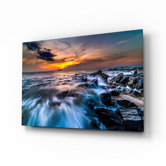 Angled view of glass wall art displaying rocky coastline with crashing waves at sunset, mounted on white background.