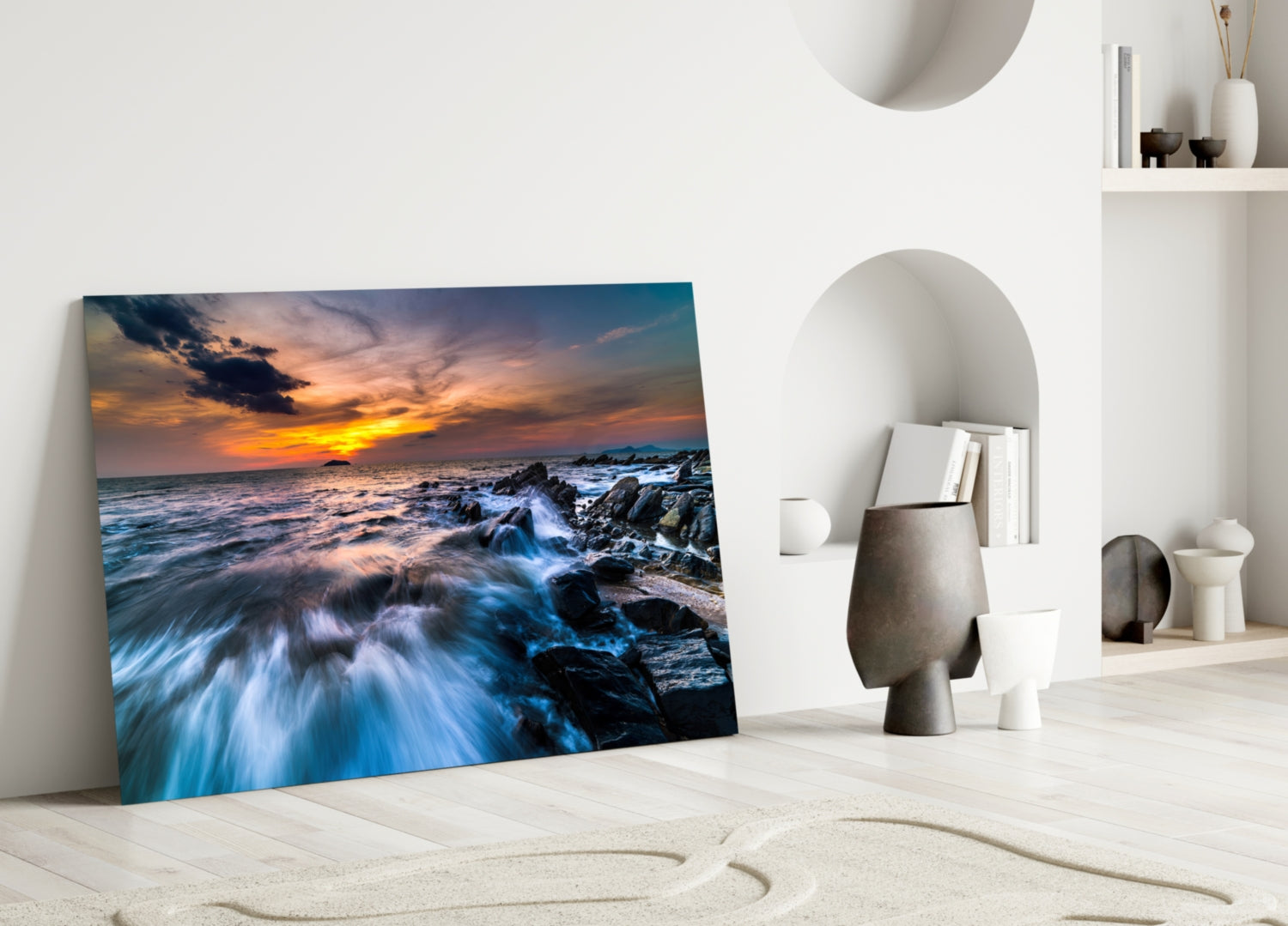 Glass wall art leaning against white wall displays rocky coastline sunset with golden sky and blue waves.