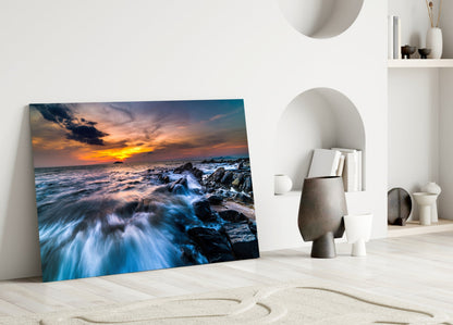 Glass wall art leaning against white wall displays rocky coastline sunset with golden sky and blue waves.