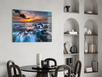 Coastal sunset photograph with orange sky and blue waves displayed on wall above round black dining table in modern interior.