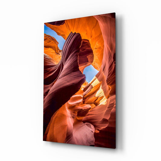 Glass wall art displaying an upward view of a slot canyon with flowing orange and red rock formations and blue sky visible through the opening.