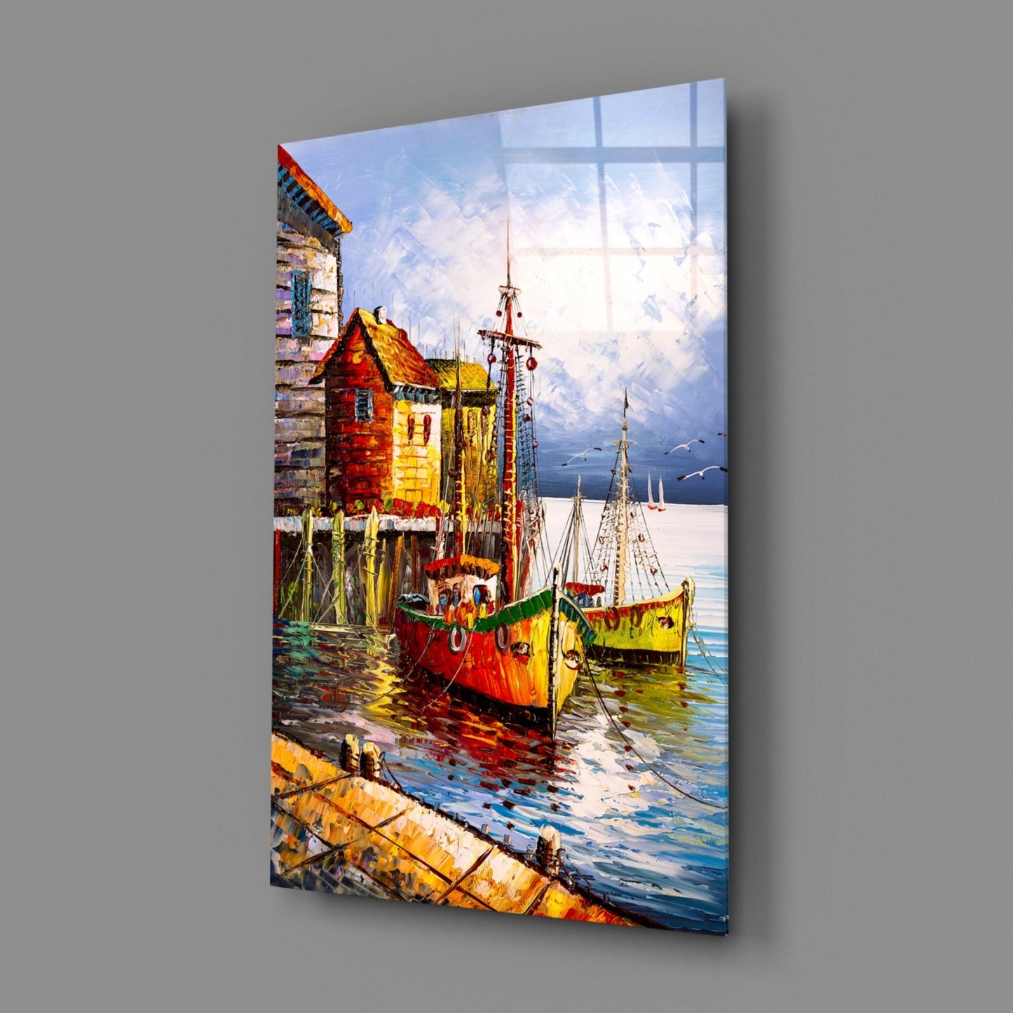 Colorful glass wall art panel displaying sailboats and waterfront cottages in vibrant reds, yellows, and blues, shown at an angle against gray background.