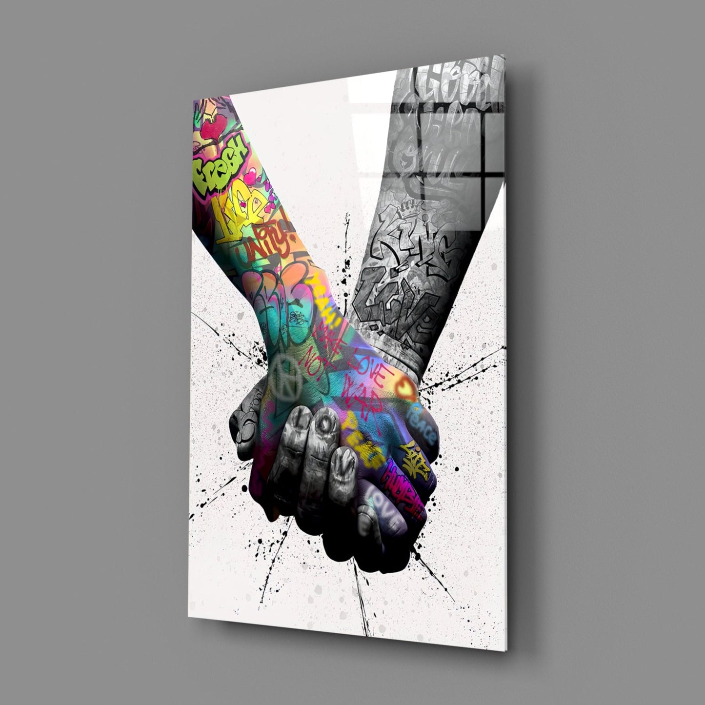Angled perspective of glass wall art featuring two hands clasped together, one colorfully graffitied, one monochrome, with ink splatter effects on white background.