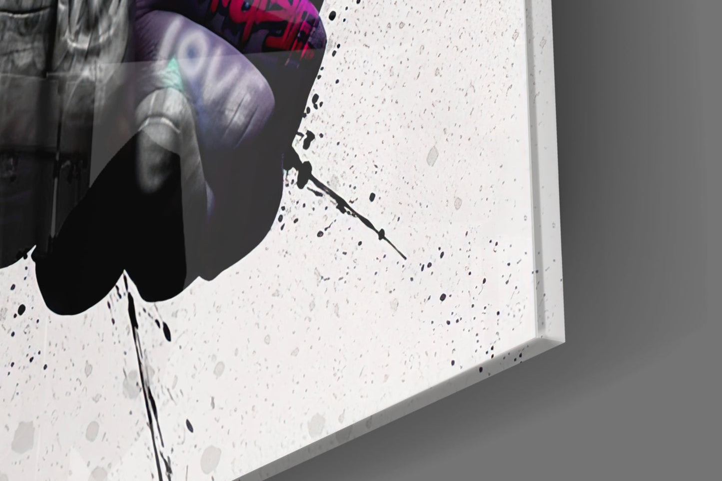 Close-up of white speckled glass wall art showing black ink splatter detail and edge profile against gray background.