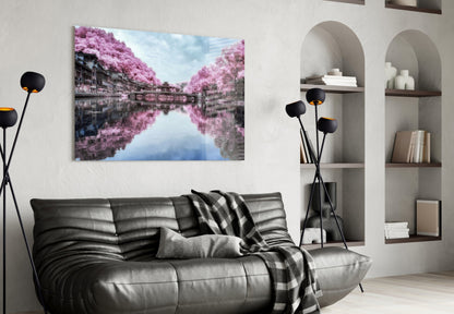 Pink landscape glass wall art displaying mirrored canal with blooming trees, hung above black leather sofa in modern living room.
