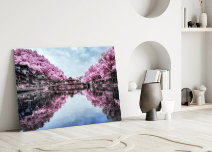 Glass wall art depicting pink flowering trees lining a canal with blue sky reflection, leaning against white wall beside decorative objects.