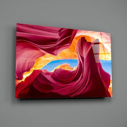 Vibrant glass wall art panel displaying layered magenta and orange canyon formations with blue sky opening, mounted on gray wall.