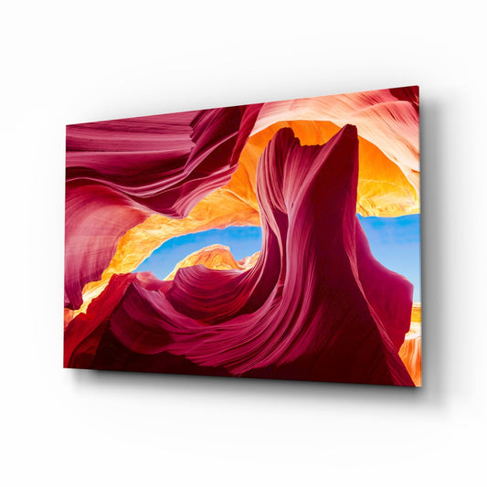Glass wall art panel displaying swirling crimson and magenta rock formations with golden and blue sky openings, angled three-quarter view.