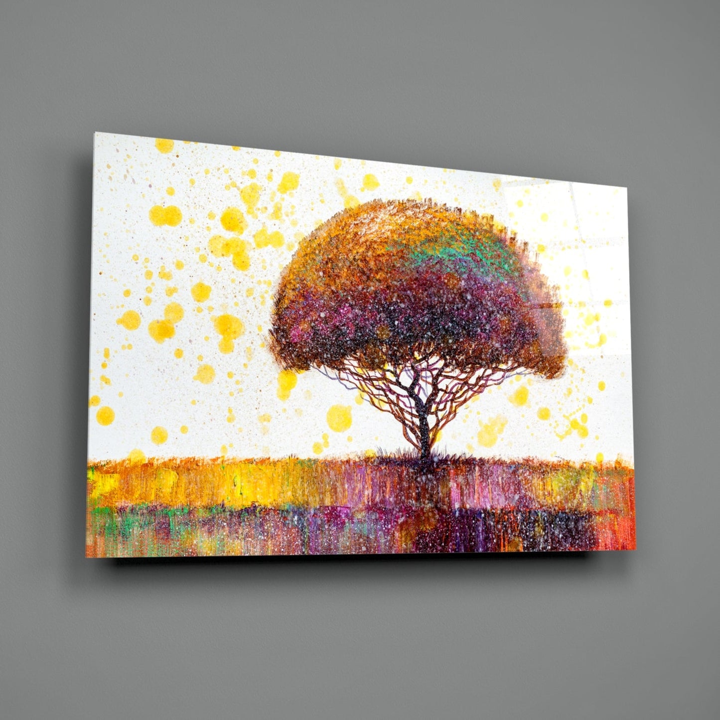 Glass wall art displaying a solitary tree with multicolored foliage against a white background with yellow paint splatters and a colorful striped ground.