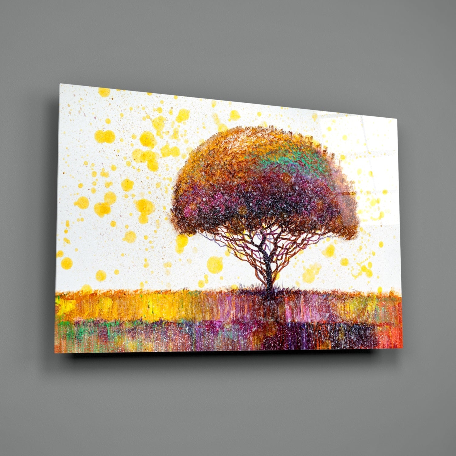 Glass wall art displaying a solitary tree with multicolored foliage against a white background with yellow paint splatters and a colorful striped ground.