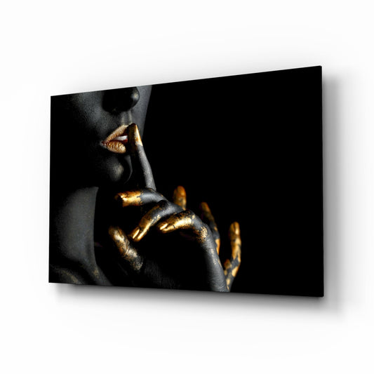 Close-up of a person's face and hand with black skin and gold metallic accents, making a shushing gesture against a black background on glass panel wall art.