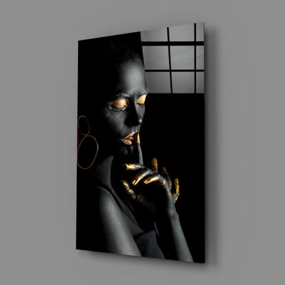 Glass wall art displaying a woman with black skin and gold accents, shown at an angle against a gray wall.