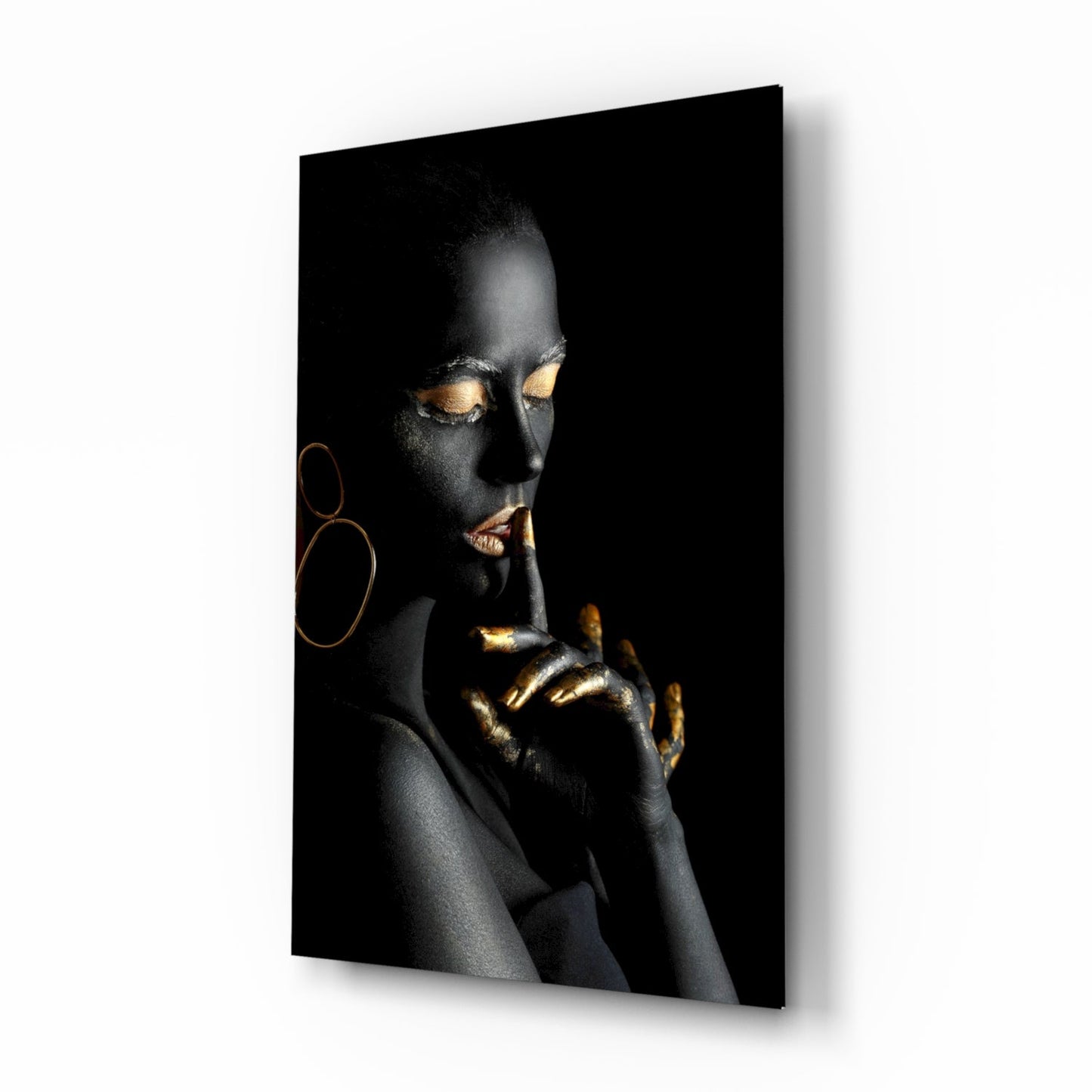 Woman with black body paint and gold leaf accents on eyes and hands against dark background, displayed as angled wall-mounted glass panel.