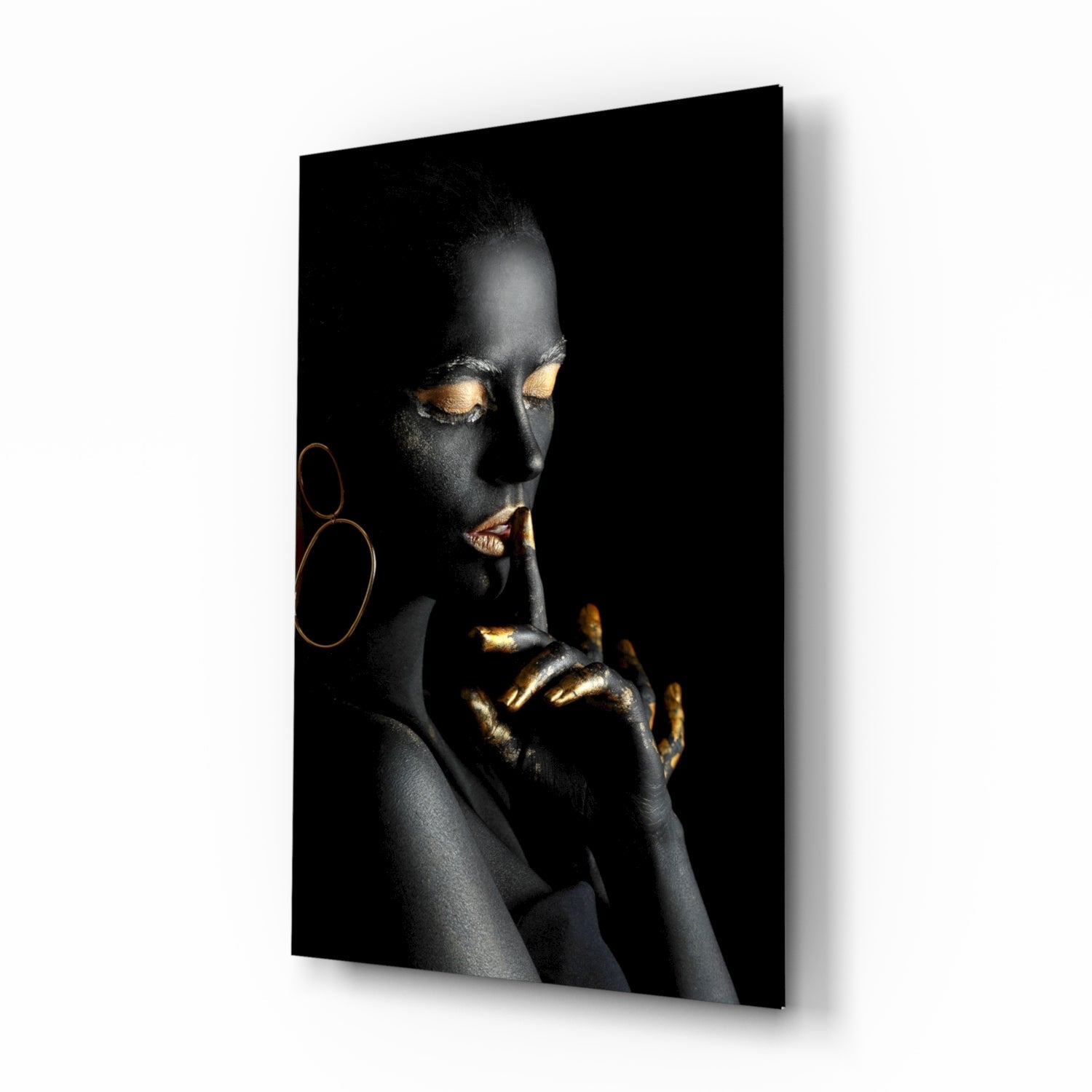 Woman with black body paint and gold leaf accents on eyes and hands against dark background, displayed as angled wall-mounted glass panel.
