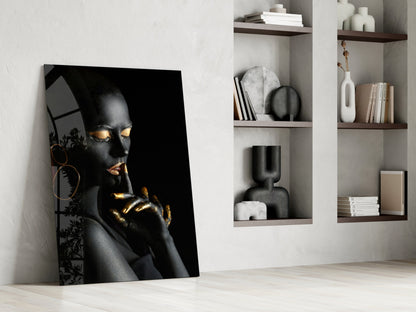 Black and gold portrait glass art leaning against white wall beside wooden shelving with decorative objects and books.