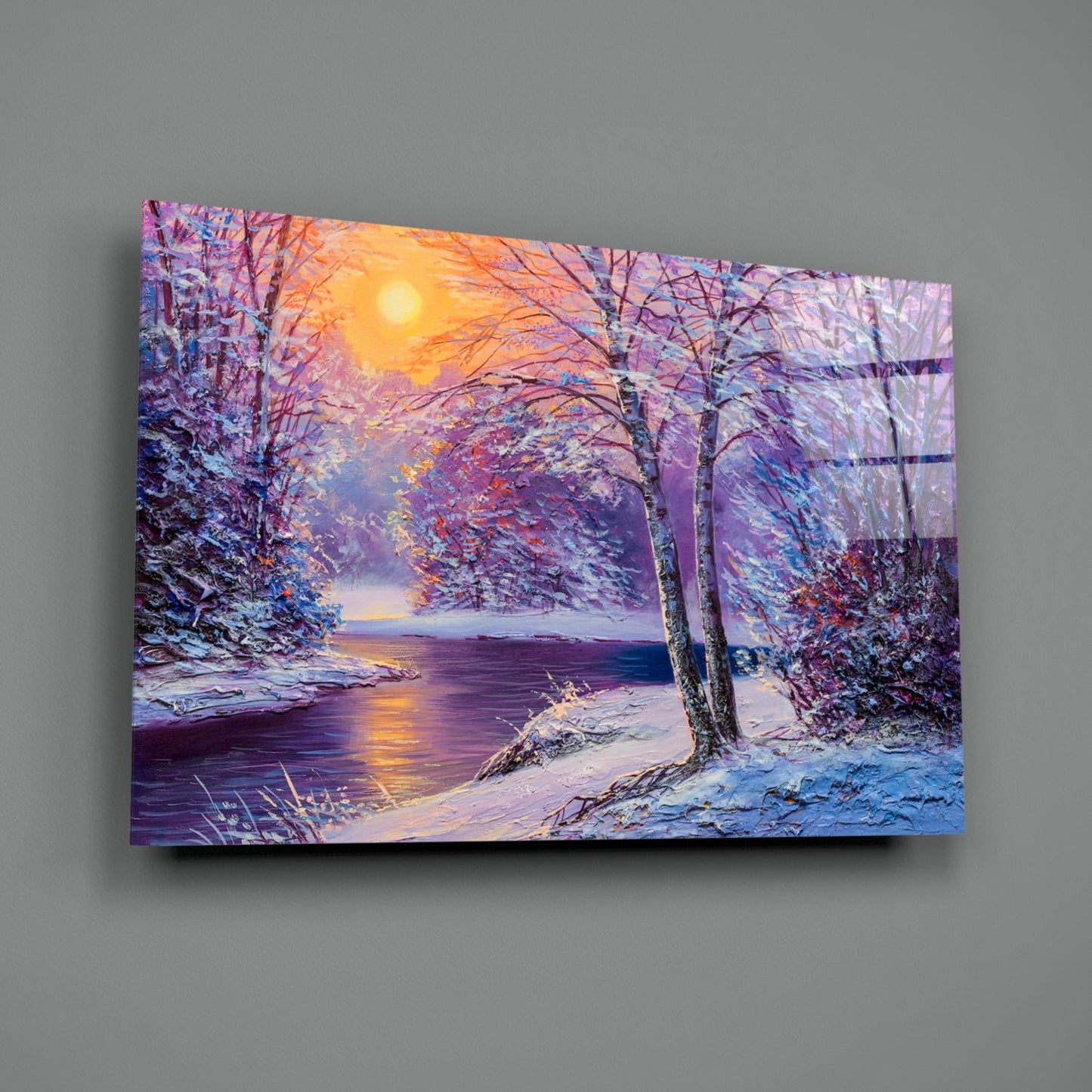 Glass wall art depicting a snow-covered riverside landscape at sunset with purple and gold tones, displayed at an angle against a gray wall.
