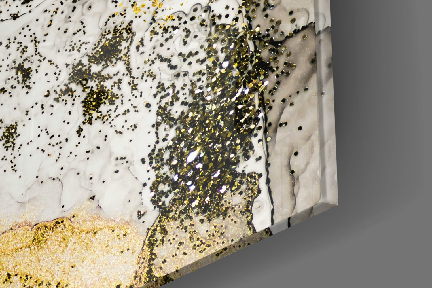 Close-up angled view of glass wall art with white resin base, gold and black glitter scattered across surface, and sandy gold texture at bottom edge.