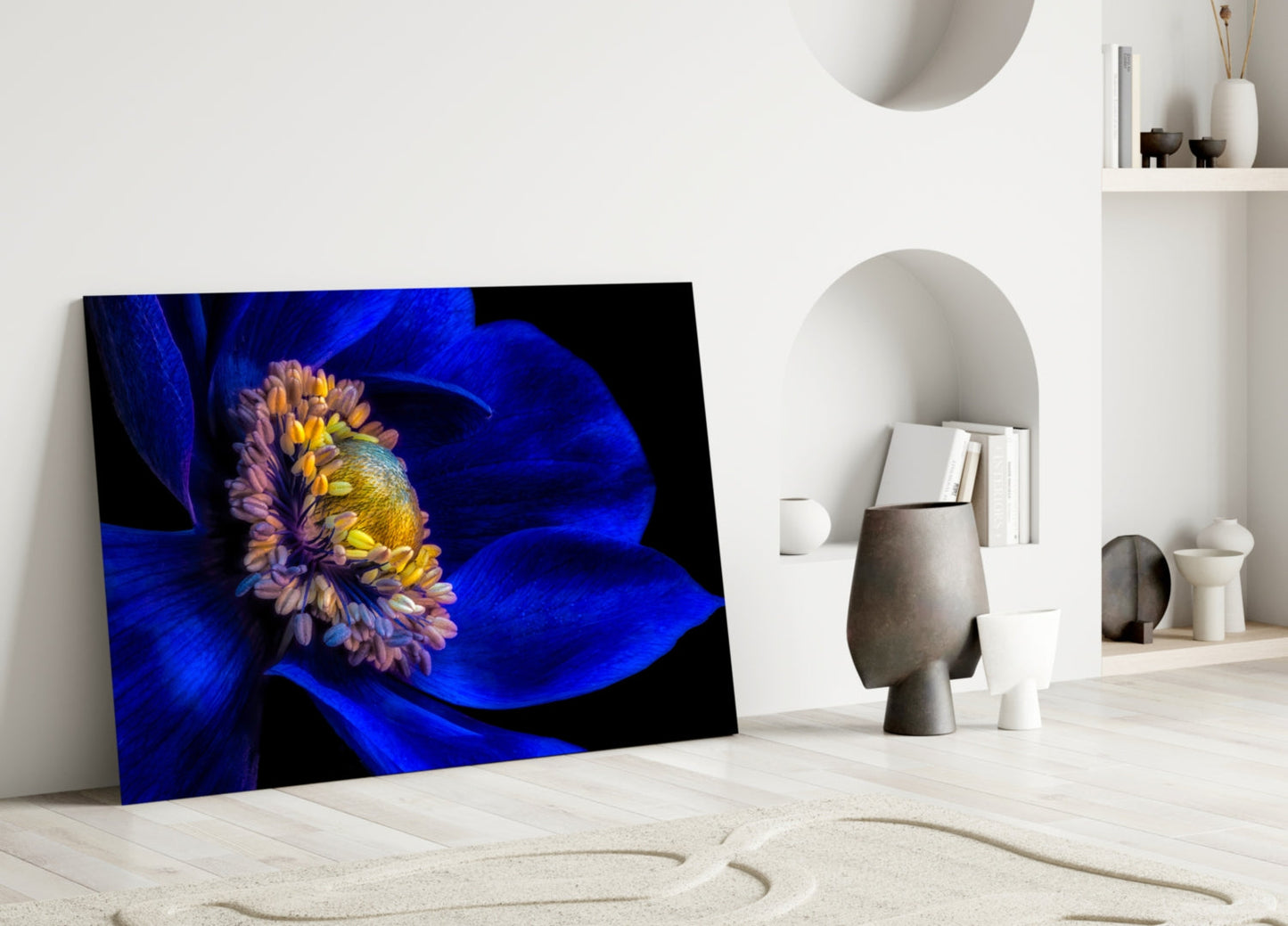 Glass wall art leaning against white wall displays vibrant blue flower with yellow center on black background in minimalist interior setting.
