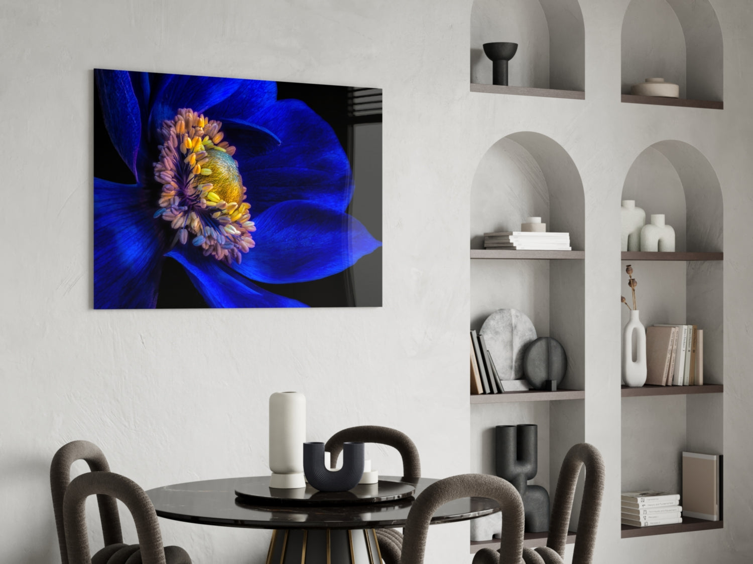 Blue peony flower with yellow center displayed as glass wall art above black dining table in modern interior setting.