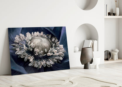 Gray flower glass wall art leaning against white wall in minimalist room setting with decorative objects and shelving.