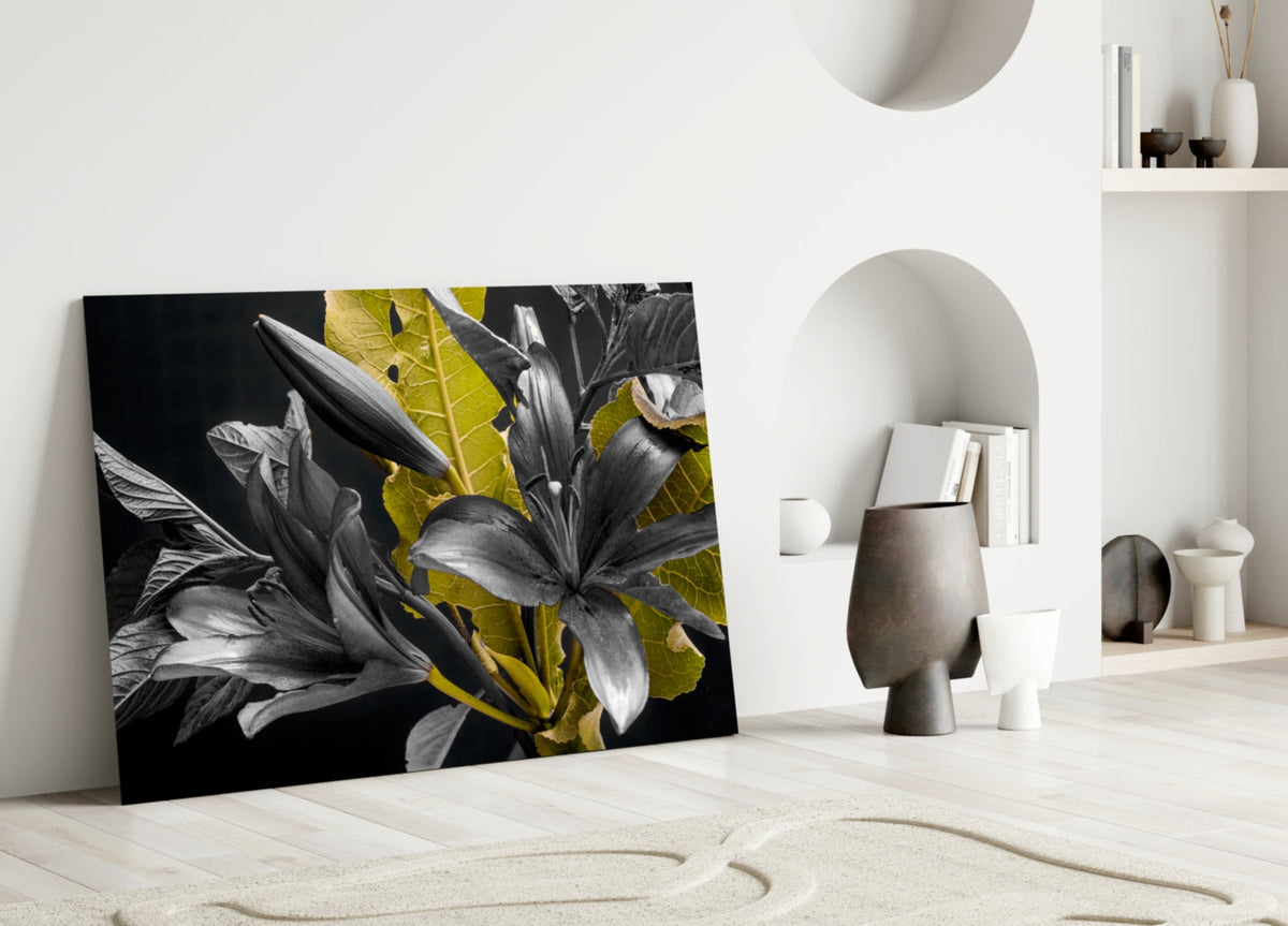 Black glass wall art featuring monochromatic lilies with golden-yellow leaves displayed on a light wooden floor in a minimalist interior setting.