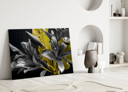 Black glass wall art featuring monochromatic lilies with golden-yellow leaves displayed on a light wooden floor in a minimalist interior setting.