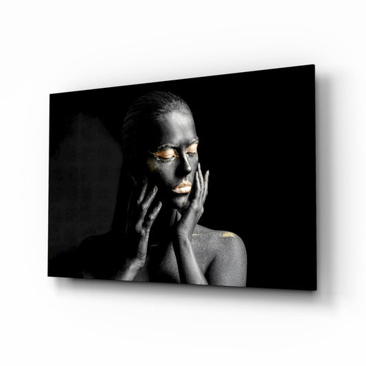 Woman with black body paint and gold eyelids and lips, hands to face, against dark background on glass wall art.