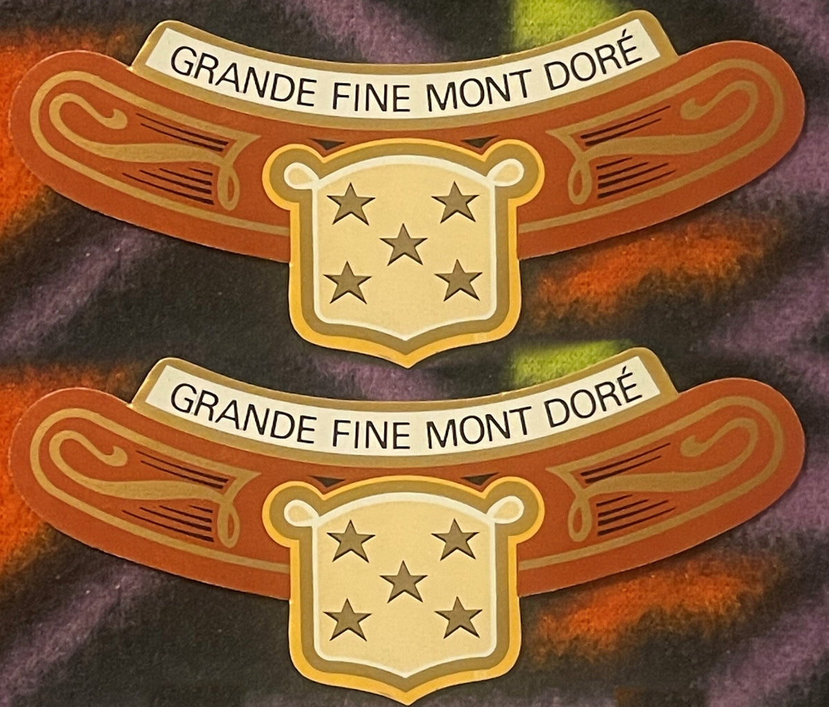 Two antique Grande Fine Mont Doré neck labels, one above the other on a colorful fleece: GRANDE FINE MONT DORÉ on a cream banner, orange arms with gold scrollwork, gold shield of five stars.