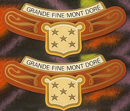 Two antique Grande Fine Mont Doré neck labels, one above the other on a colorful fleece: GRANDE FINE MONT DORÉ on a cream banner, orange arms with gold scrollwork, gold shield of five stars.