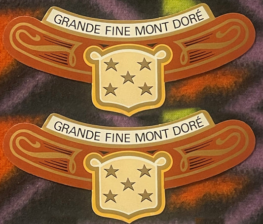 Two antique Grande Fine Mont Doré neck labels, one above the other on a colorful fleece: GRANDE FINE MONT DORÉ on a cream banner, orange arms with gold scrollwork, gold shield of five stars.