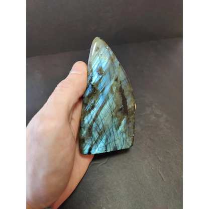 A hand holds a polished labradorite free form sculpture with iridescent blue and green flashes on a triangular stone