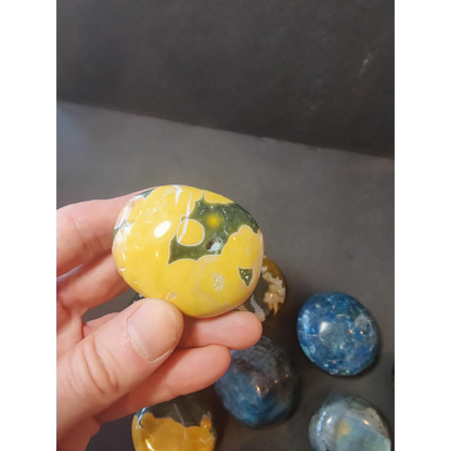 A smooth oval-shaped yellow and green marbled polished palmstone held in a hand for assorted stone collection