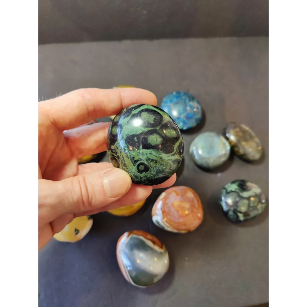 A hand holds a polished palmstone green and black agate with swirling eye-like formations assorted stones