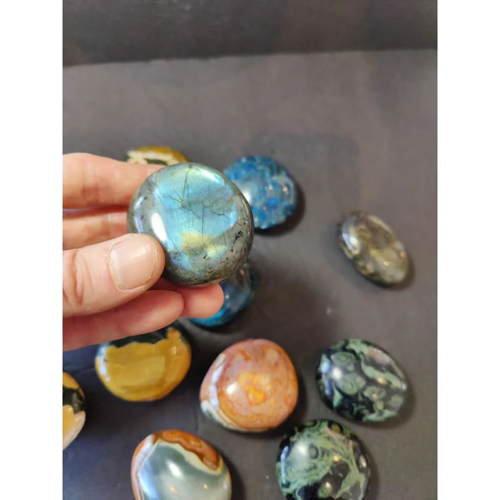 A hand holds a polished palmstone labradorite with iridescent blue and green hues shimmering under light