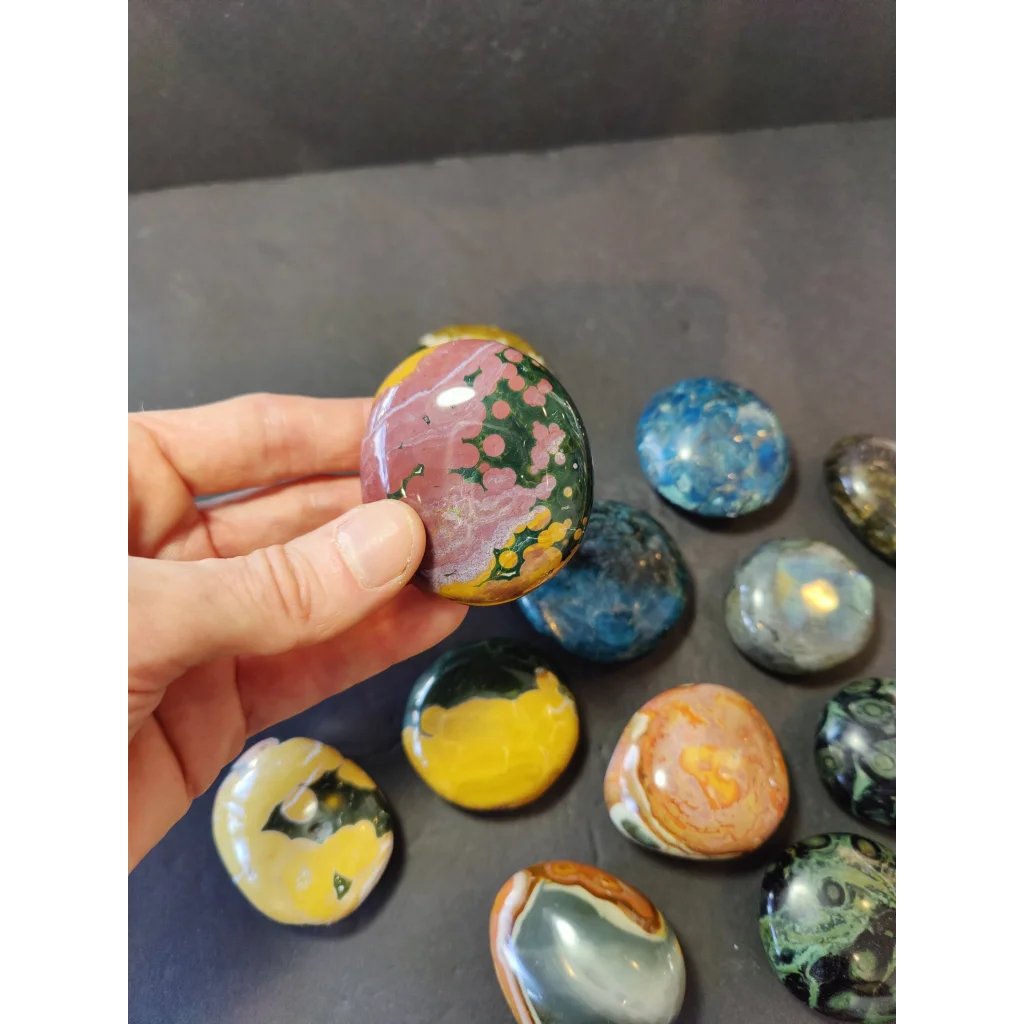 A hand holds a polished jasper palmstone with swirling pink and green patterns among assorted colorful stones