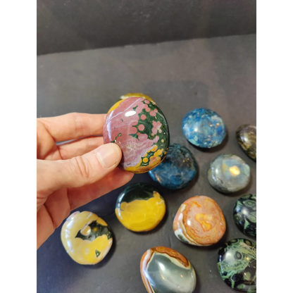 A hand holds a polished jasper palmstone with swirling pink and green patterns among assorted colorful stones