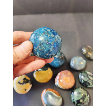 A hand holds a polished blue agate palmstone sphere with intricate crackled patterns and natural mineral inclusions