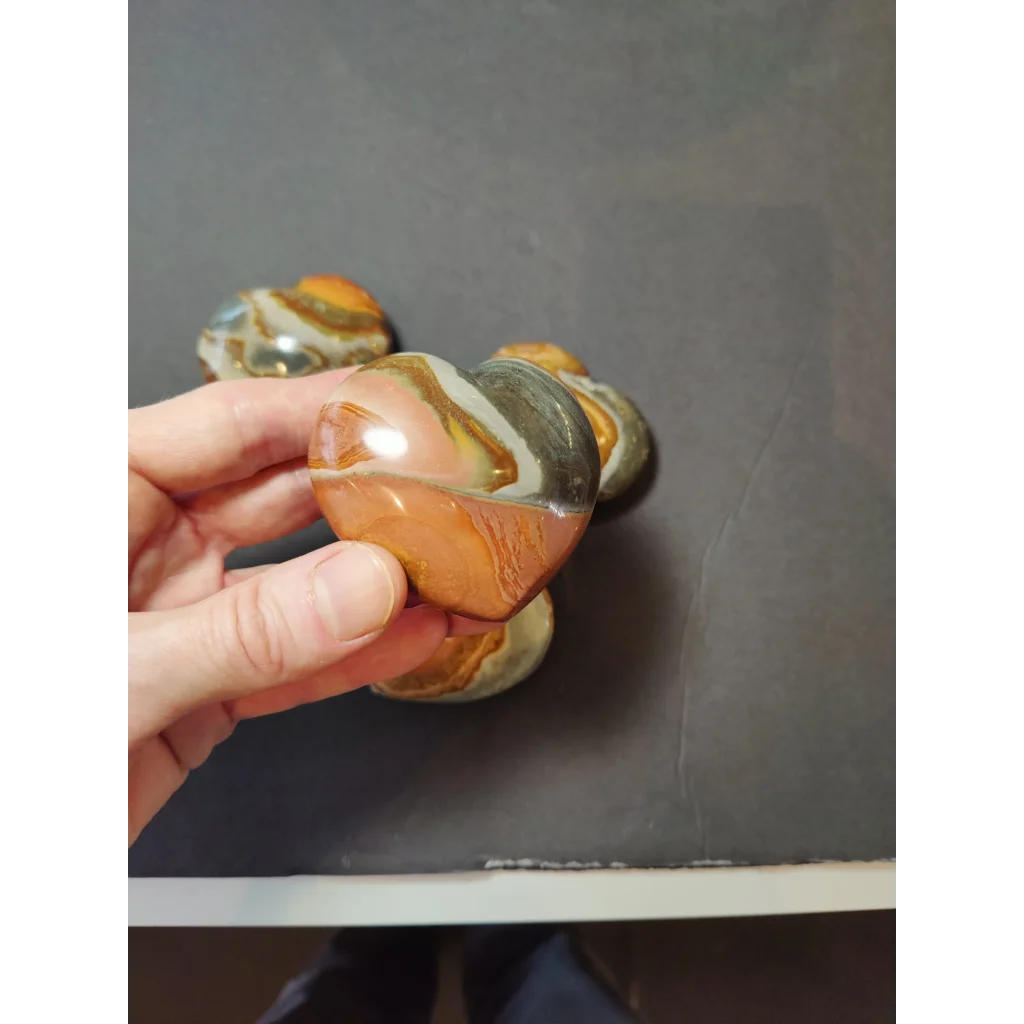 A hand holds a polished heart-shaped polychrome jasper carved stone with swirling orange brown and white bands