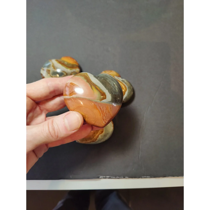 A hand holds a polished heart-shaped polychrome jasper carved stone with swirling orange brown and white bands