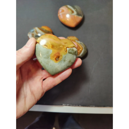 A hand holds a polished heart-shaped polychrome jasper carved stone with orange and green swirling patterns