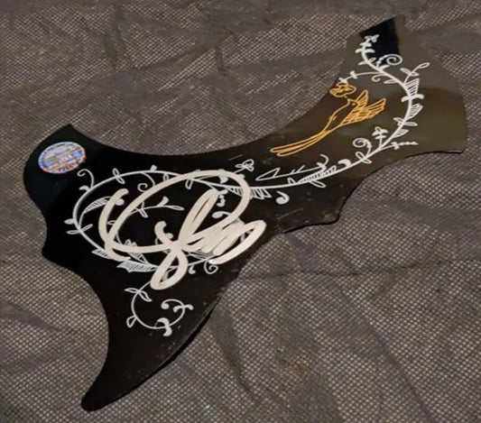 Black guitar pickguard with white and gold floral vine designs and gold bird, displayed flat on gray textile background.