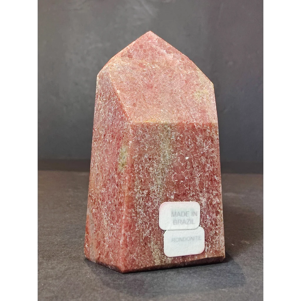 A polished reddish-pink rhodochrosite polished obelisk with a textured crystalline surface and two white labels at its base