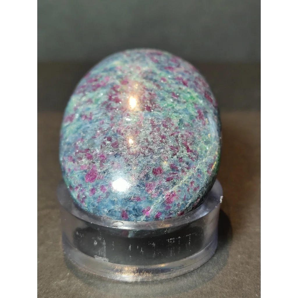 A polished oval-shaped ruby kyanite stone with teal and pink speckles displayed on a clear acrylic stand