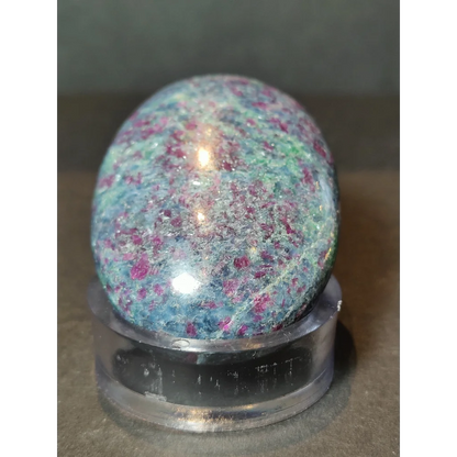 A polished oval-shaped ruby kyanite stone with teal and pink speckles displayed on a clear acrylic stand
