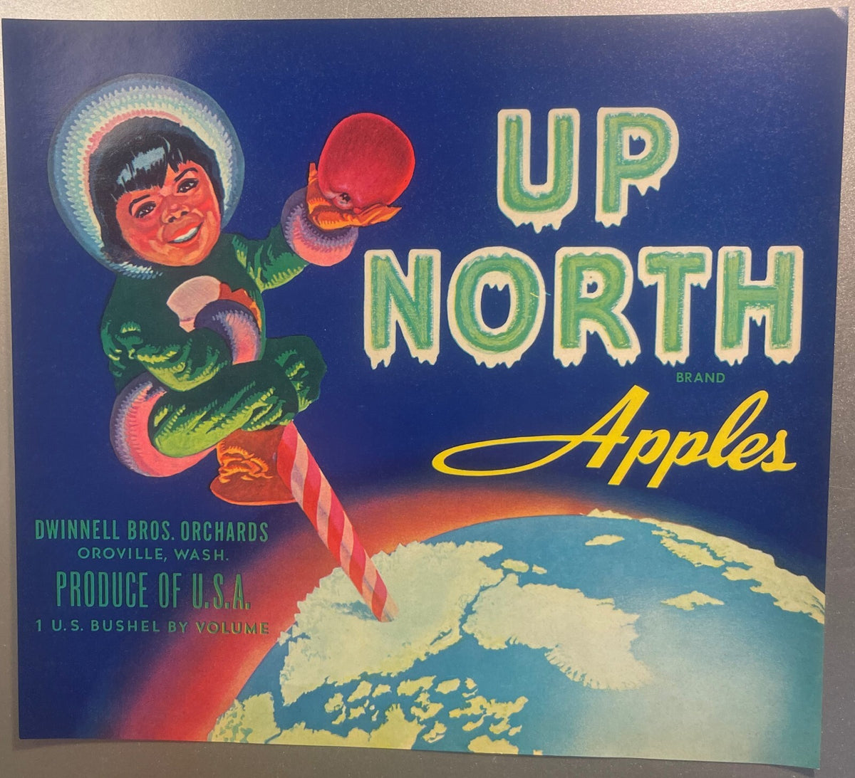 Vintage 1940s Up North Brand Apples crate label from Dwinnell Bros. Orchards, Oroville, Wash.: a smiling Inuit child holds up a red apple on a candy-striped North Pole at the top of the globe, icicle lettering on deep blue
