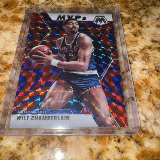 A 2019-20 Mosaic MVPs basketball card featuring Wilt Chamberlain in a Philadelphia 76ers uniform, displaying a holographic blue prizm background with colorful geometric patterns, photographed flat on a speckled granite surface.