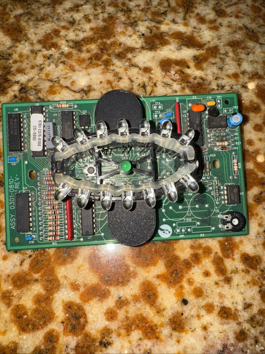 Top-down view of green circuit board with transparent plastic dome cover and silver metal components mounted on speckled granite countertop.